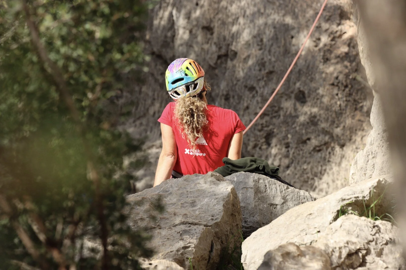 A woman with curly hair wearing a colorful helmet and red T-shirt sitting on a rock, looking at a rock climbing wall with a safety rope attached.