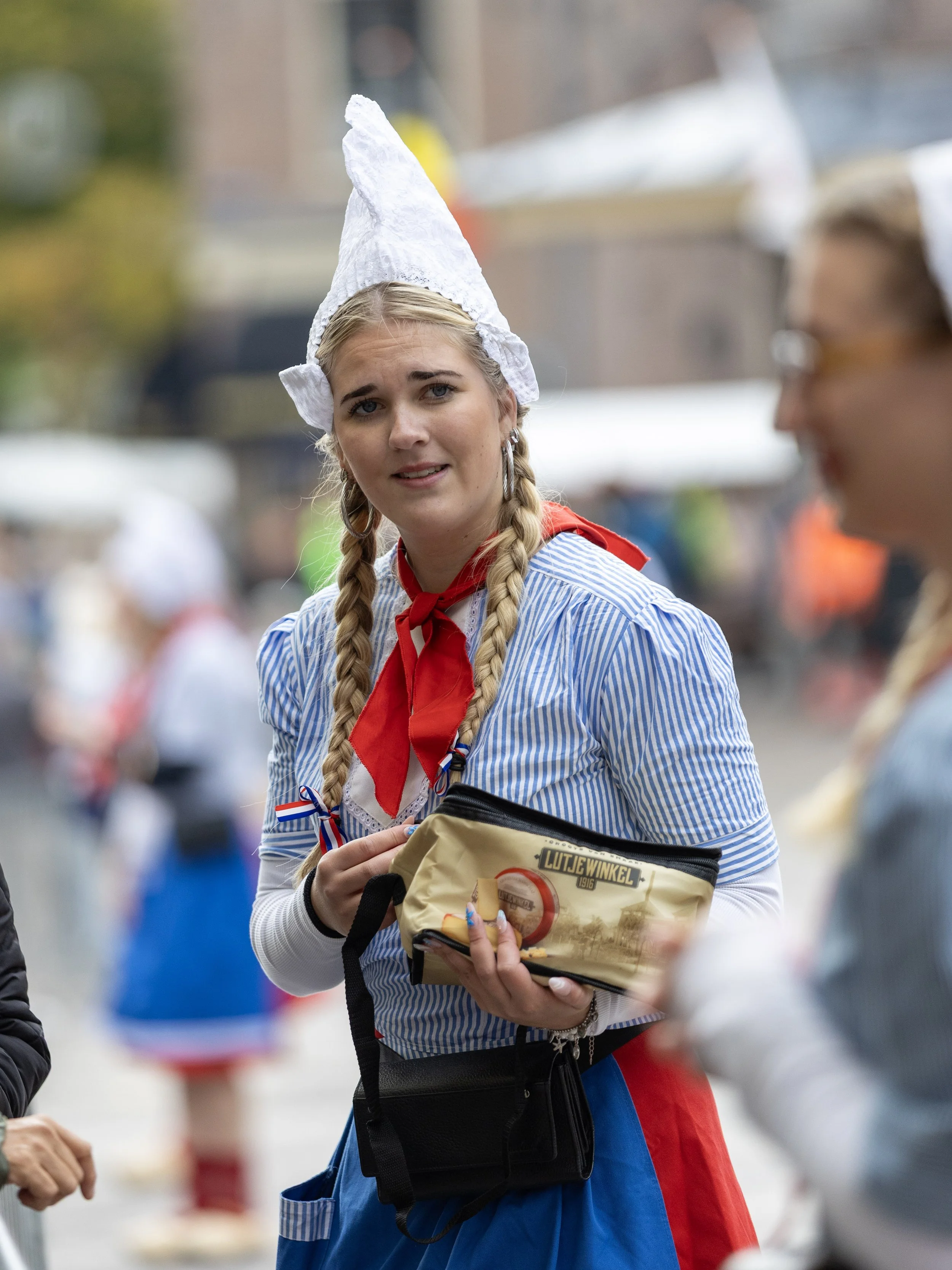 A young woman dressed in traditional Dutch attire with pigtails, a white bonnet, and a striped shirt, holding a beige bag and engaged in conversation at a festival or parade.