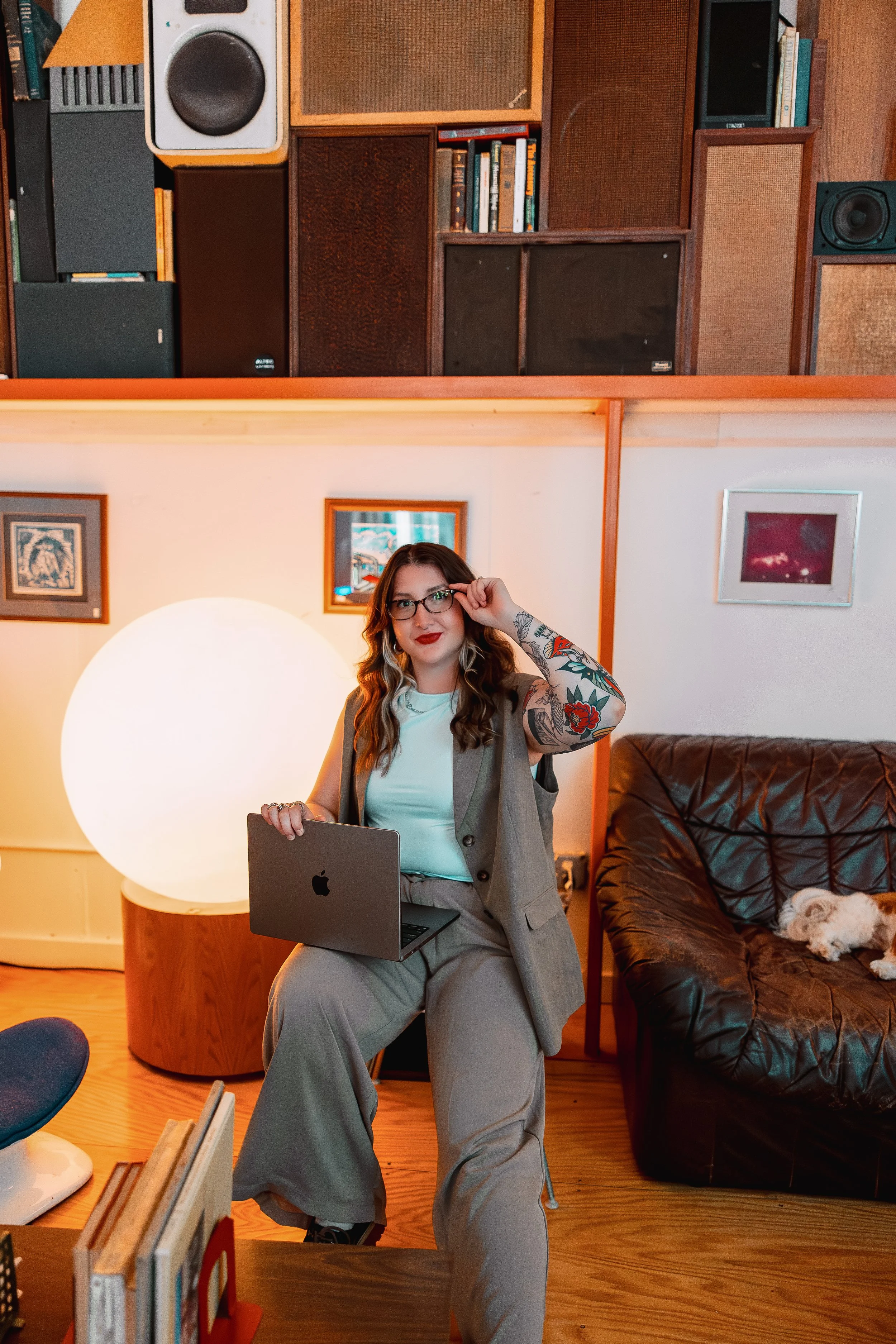 A woman with glasses and tattoos is sitting on a chair, holding an Apple MacBook laptop. She has red lipstick and wavy brown hair. Behind her, there is a large round white lamp and a couch with a small dog resting on it. The room has wooden floors an