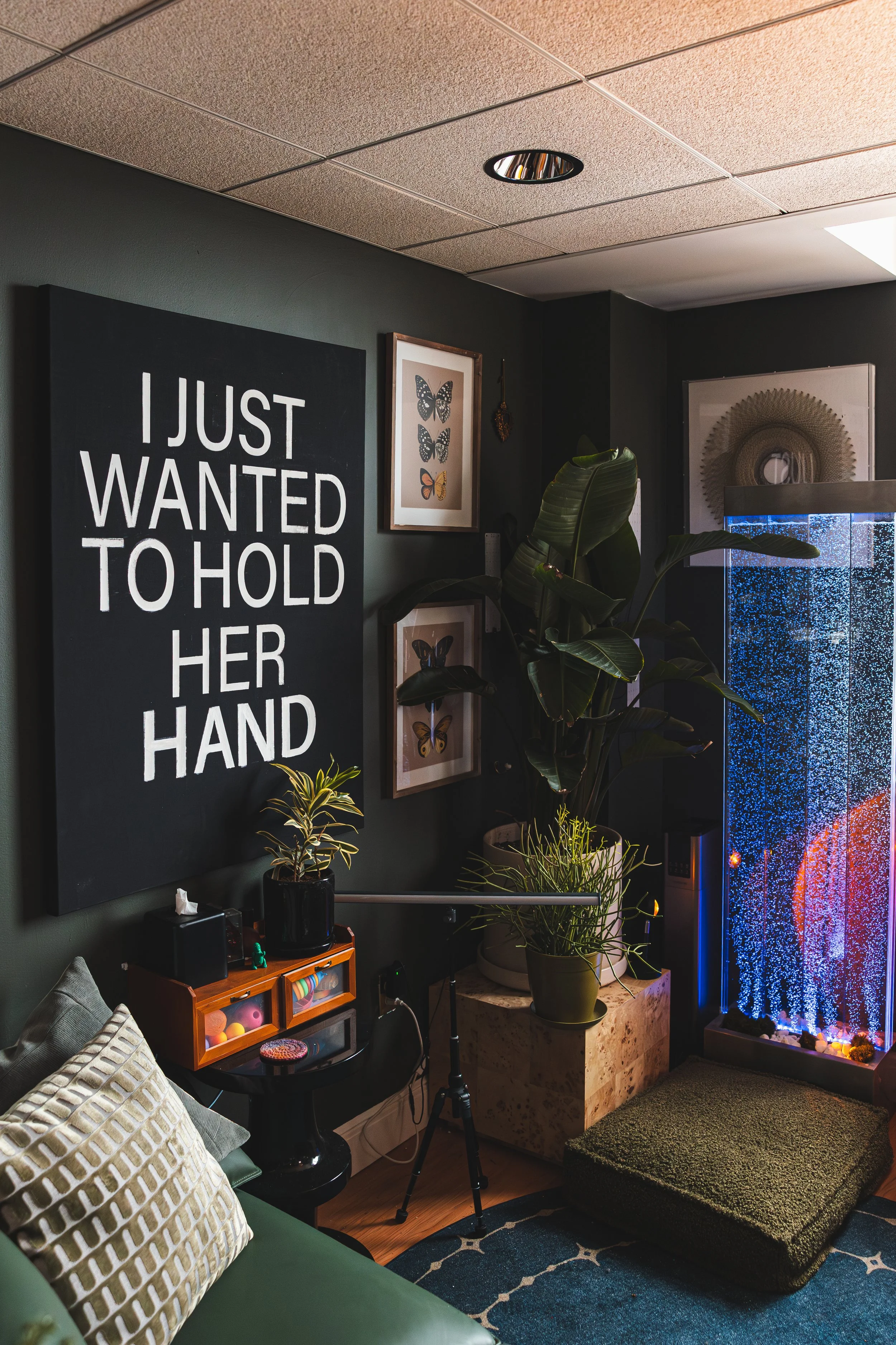 A cozy corner with a dark wall decorated with framed butterfly artwork and a large black and white sign that reads, "I just wanted to hold her hand." There are several green potted plants, a black side table with a small potted plant, a green sofa wi