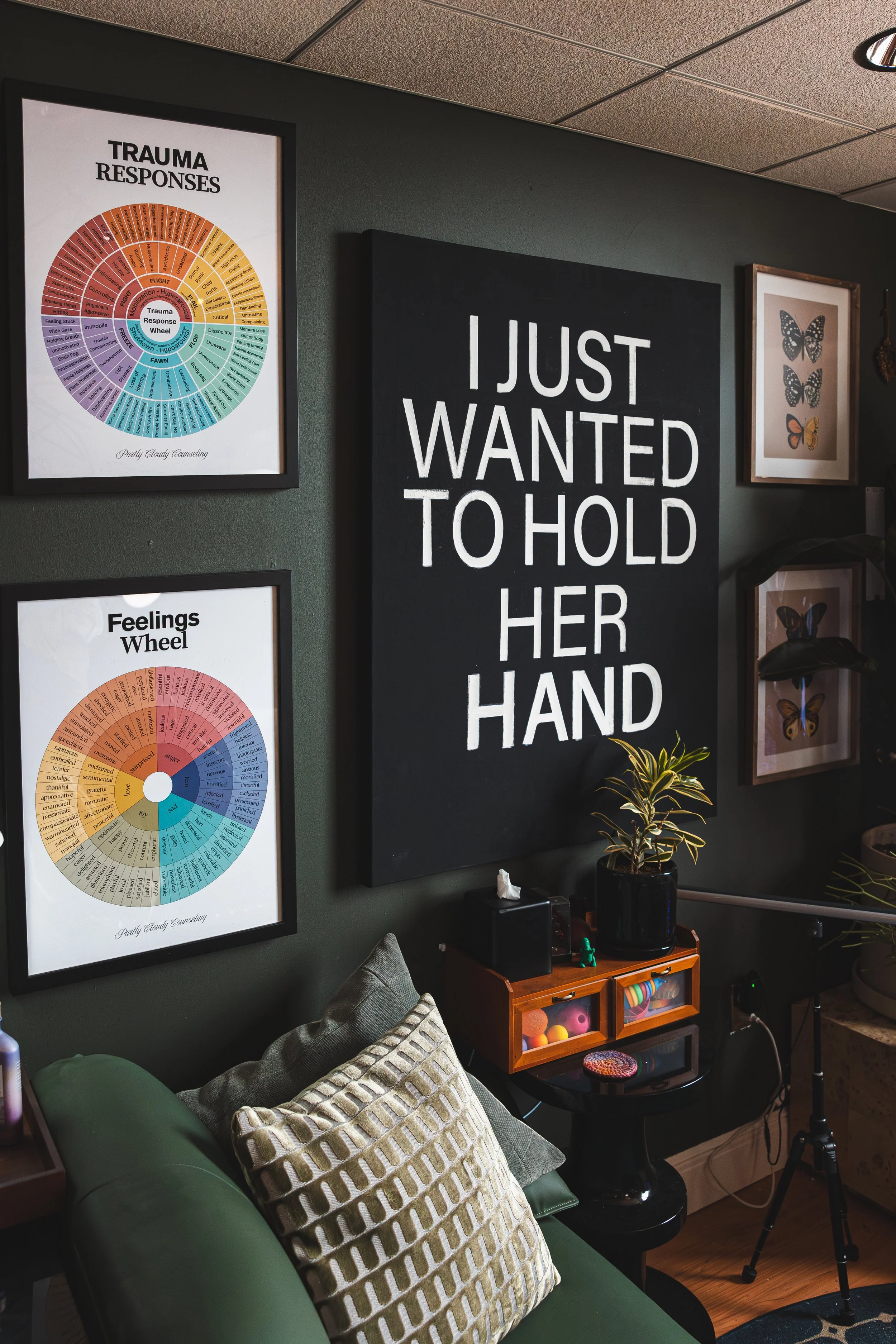 A cozy counseling office wall decorated with butterfly artwork, trauma response and feelings wheel charts, a large black sign that says "I just wanted to hold her hand," a green sofa with patterned pillows, and a small wooden cabinet with plants and