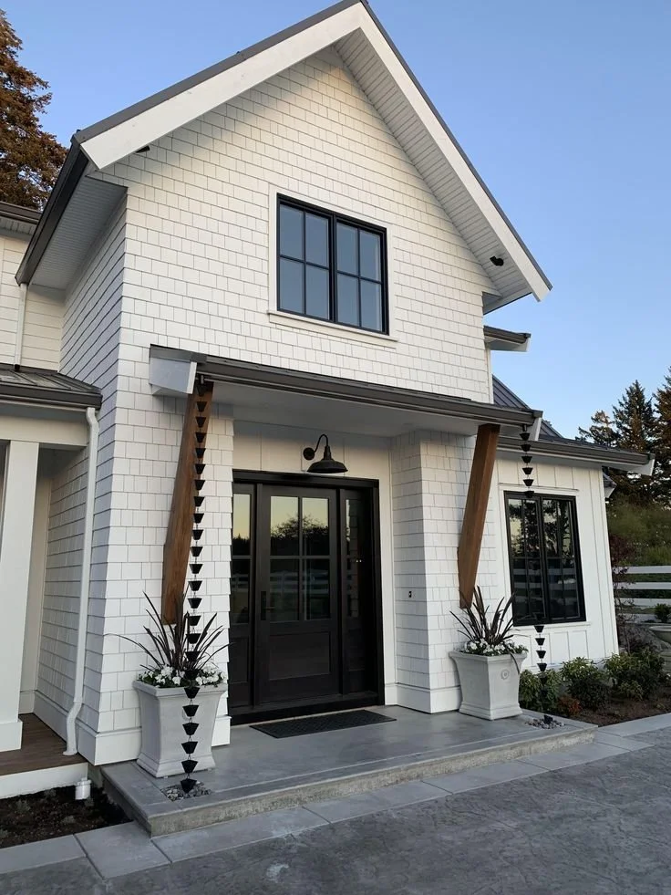 Modern white two-story house with black front door, large window, decorative planters, and wooden accents.
