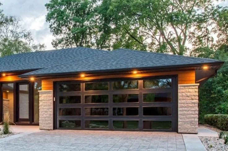 Modern house with a blue shingled roof, brick columns, and a black garage door, surrounded by greenery.