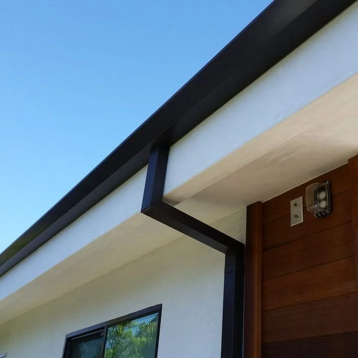 Close-up of the corner of a modern house roof with black trim, white walls, wooden siding, black modern gutters, a small outdoor light, and a window, against a clear blue sky.
