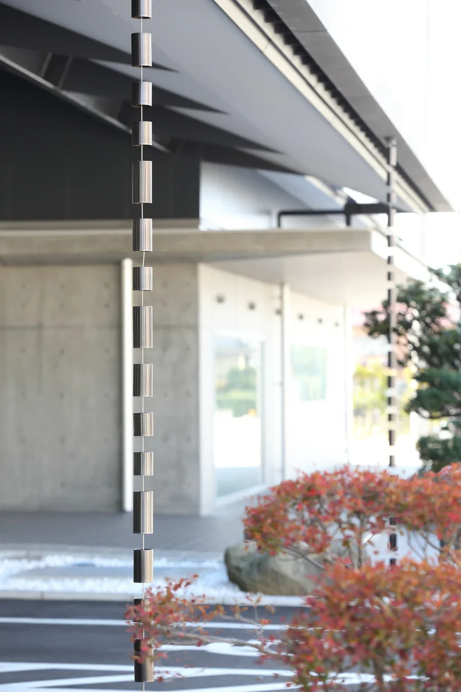 Close-up of a modern hanging air or wind chime with small metal cylinders, in front of a contemporary building with large glass windows and a tree with red and green leaves.