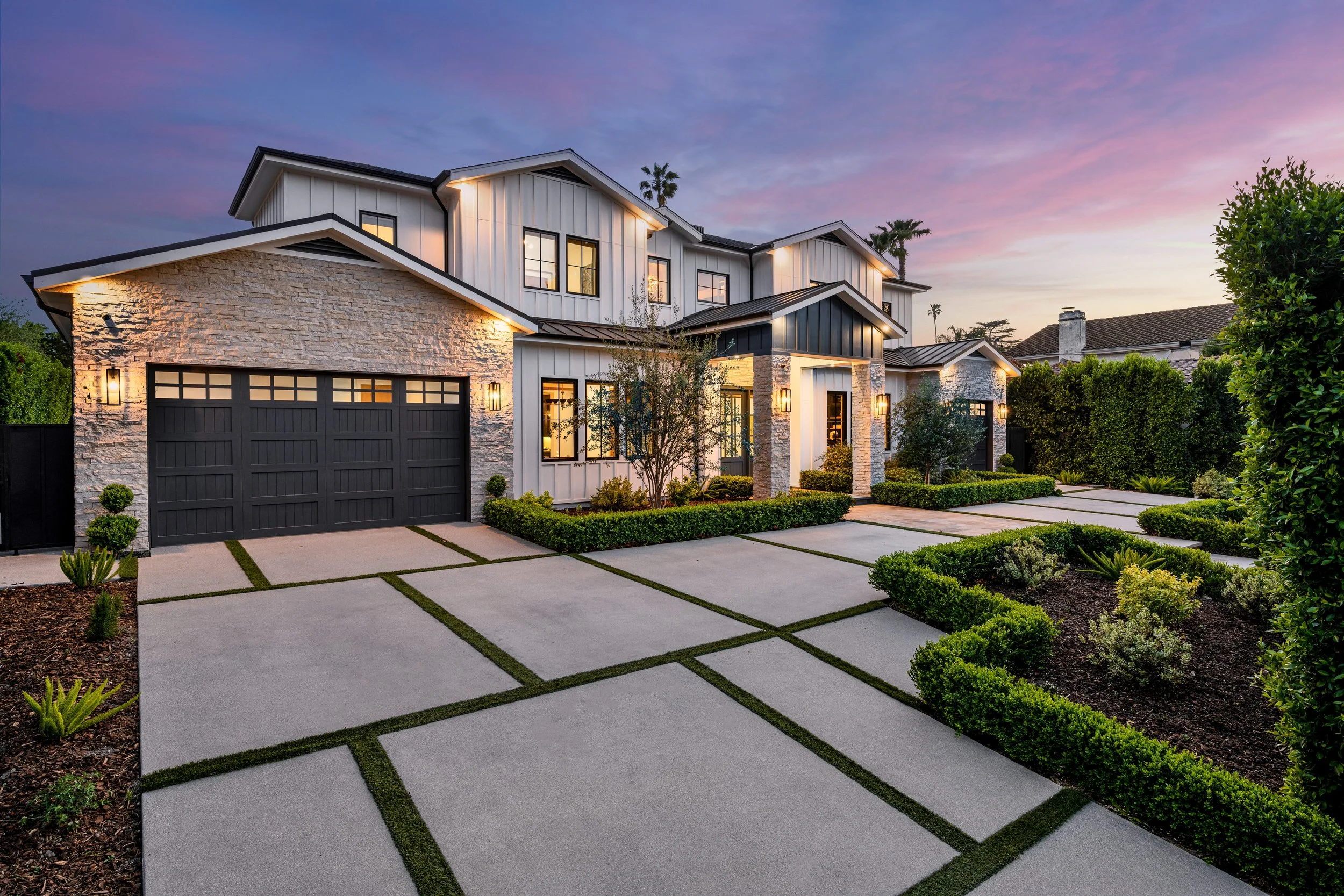 Modern two-story house with a black garage door, white exterior walls, black window frames, lush landscaping, and a spacious concrete driveway at dusk with a colorful sky.