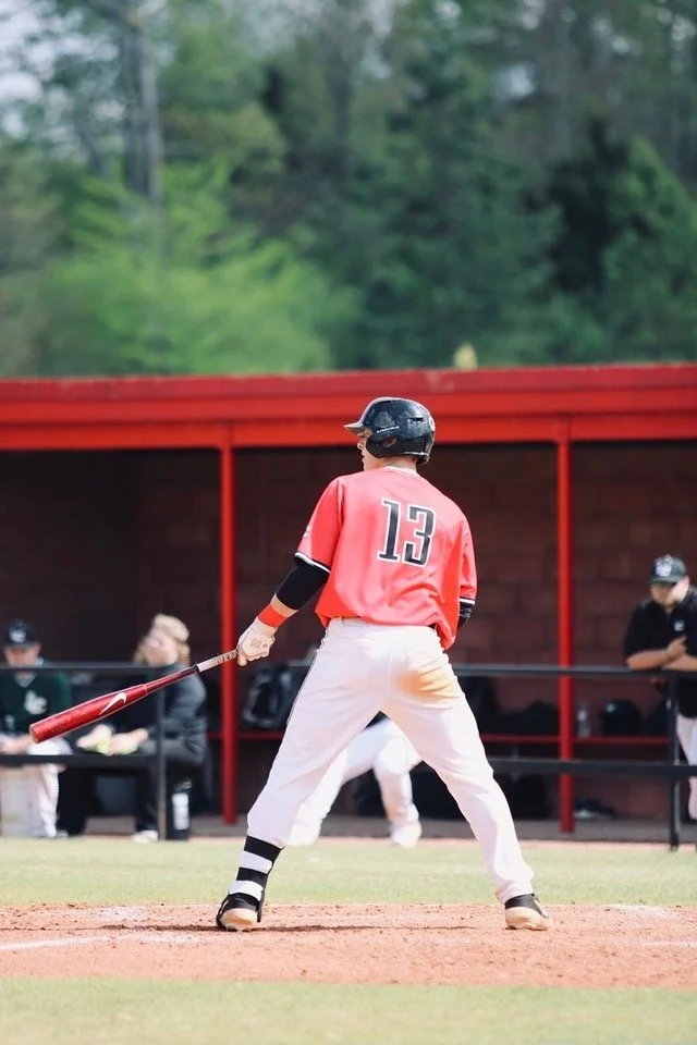 Baseball player wearing a red jersey with the number 13, black helmet, and white pants holding a bat on the field during a game.