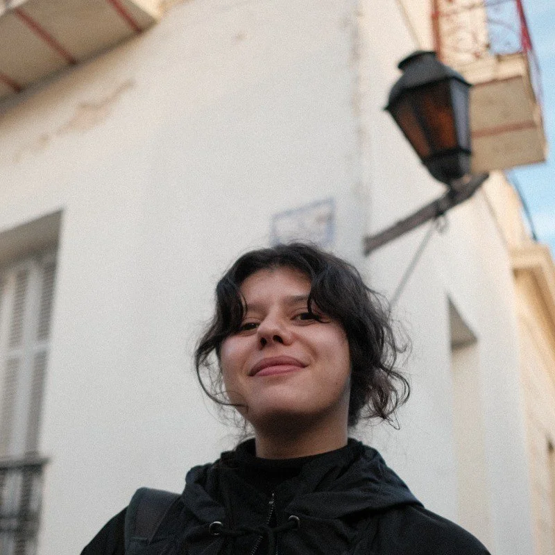 Pessoa com cabelo cacheado escuro sorrindo em rua com prédio branco ao fundo