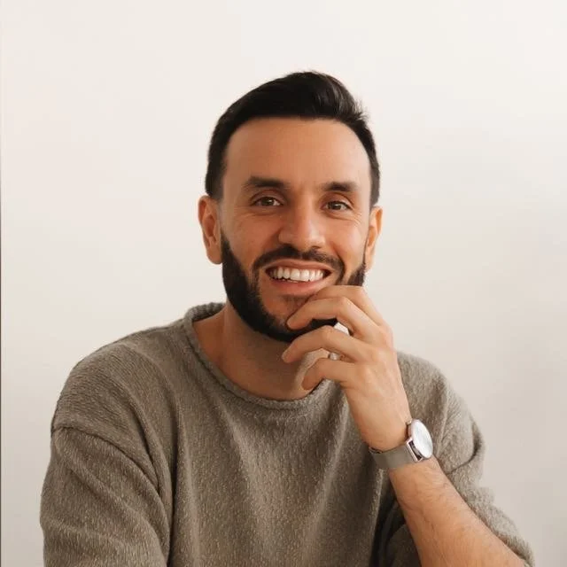 Homem sorrindo, usando suéter cinza e relógio, com cabelo escuro e barba, contra fundo branco.