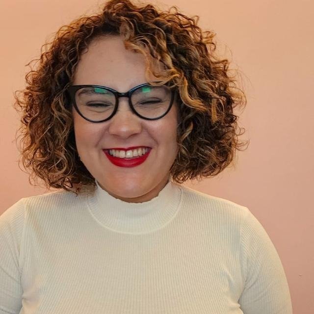 Mulher com cabelos cacheados, óculos redondos e sorriso, usando blusa branca, contra parede rosa.