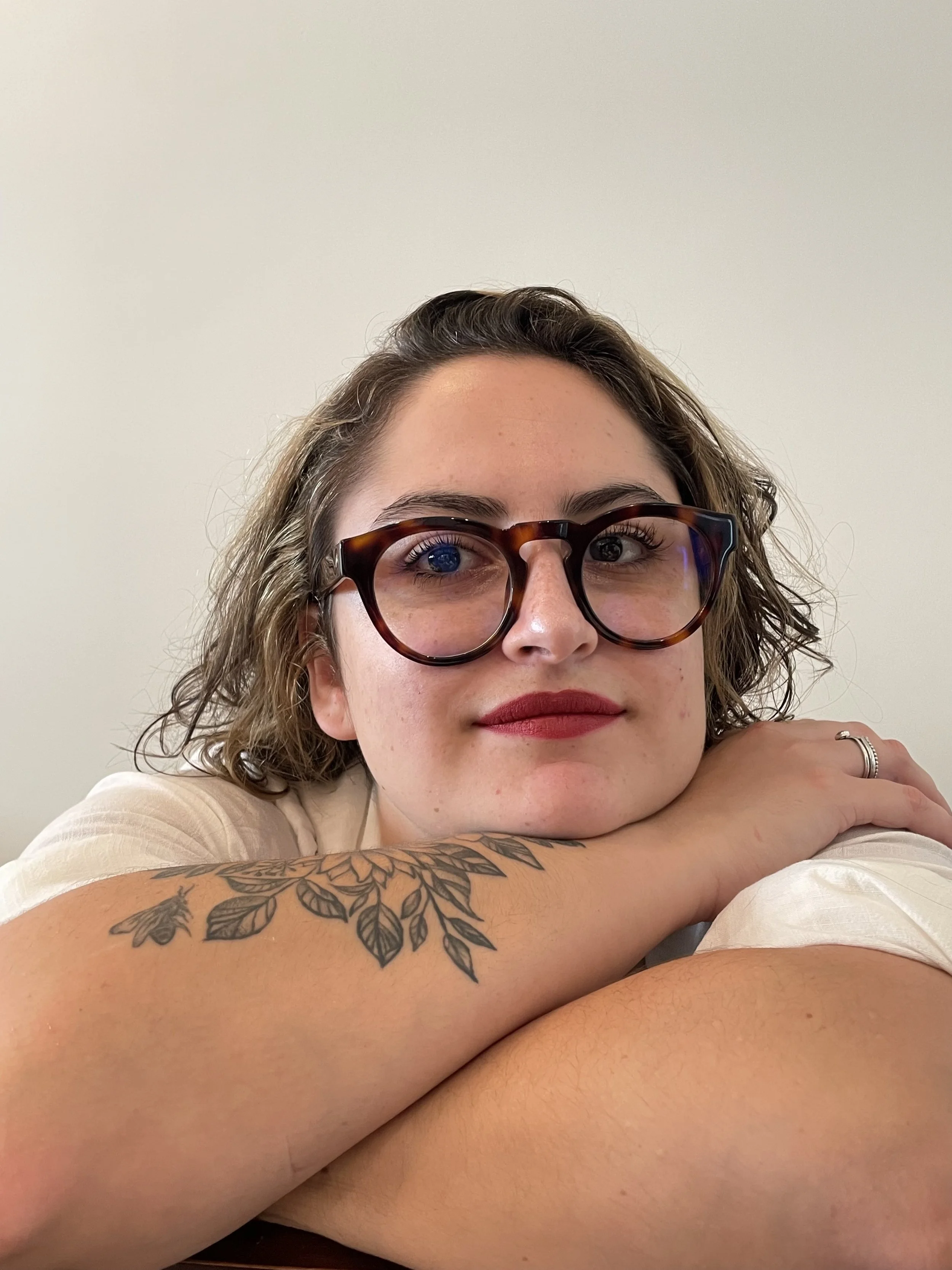 Mulher com óculos de armação grossa, cabelo ondulado, com tatuagem de folhas no braço, olhando para a câmera com expressão tranquila, apoiando a cabeça na mão sobre o braço, contra fundo branco.