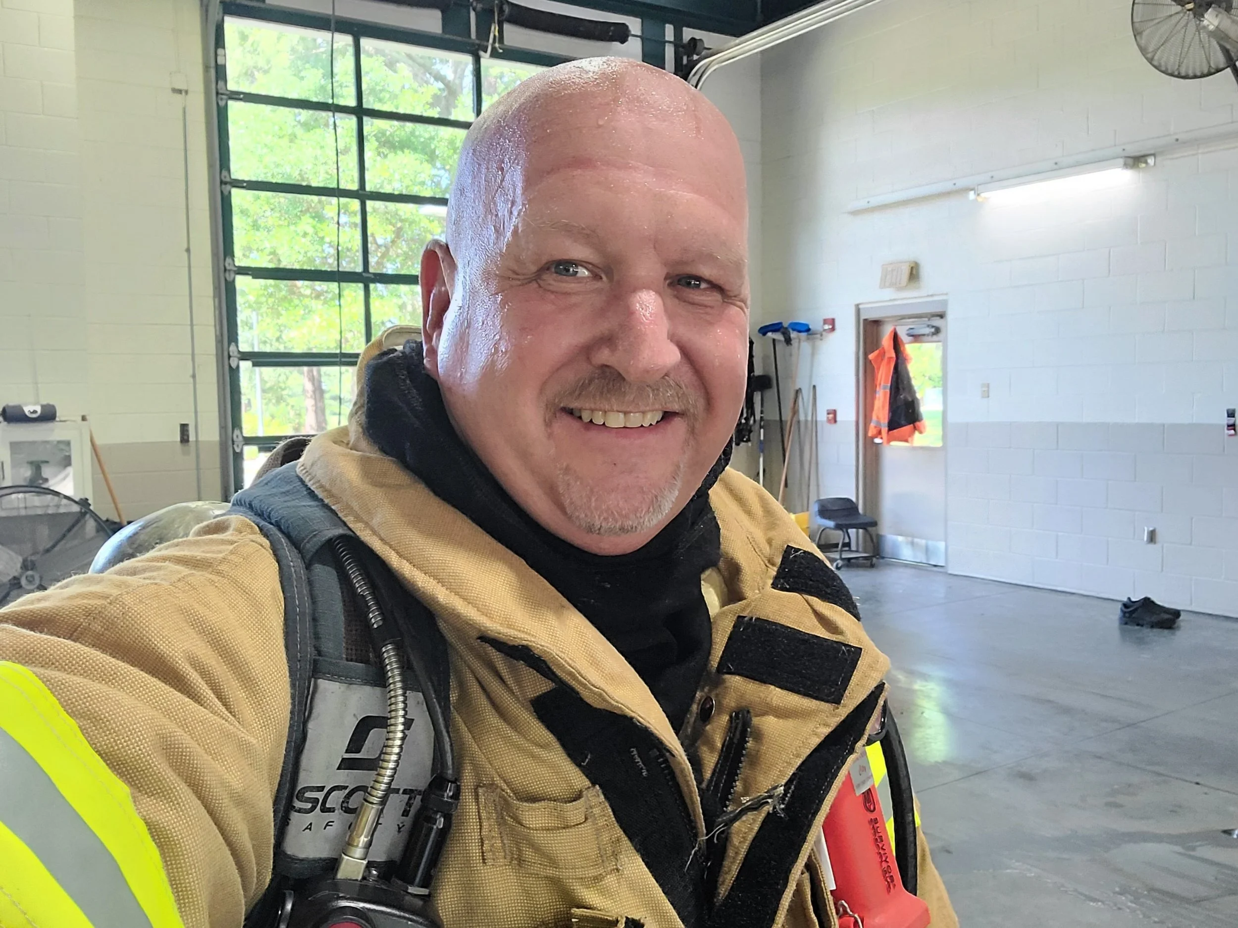 A firefighter smiling indoors in a fire station, wearing a tan turnout coat with black accents, a black undershirt, and gear on his shoulder. The background shows a large open garage with windows, a door, and various equipment.