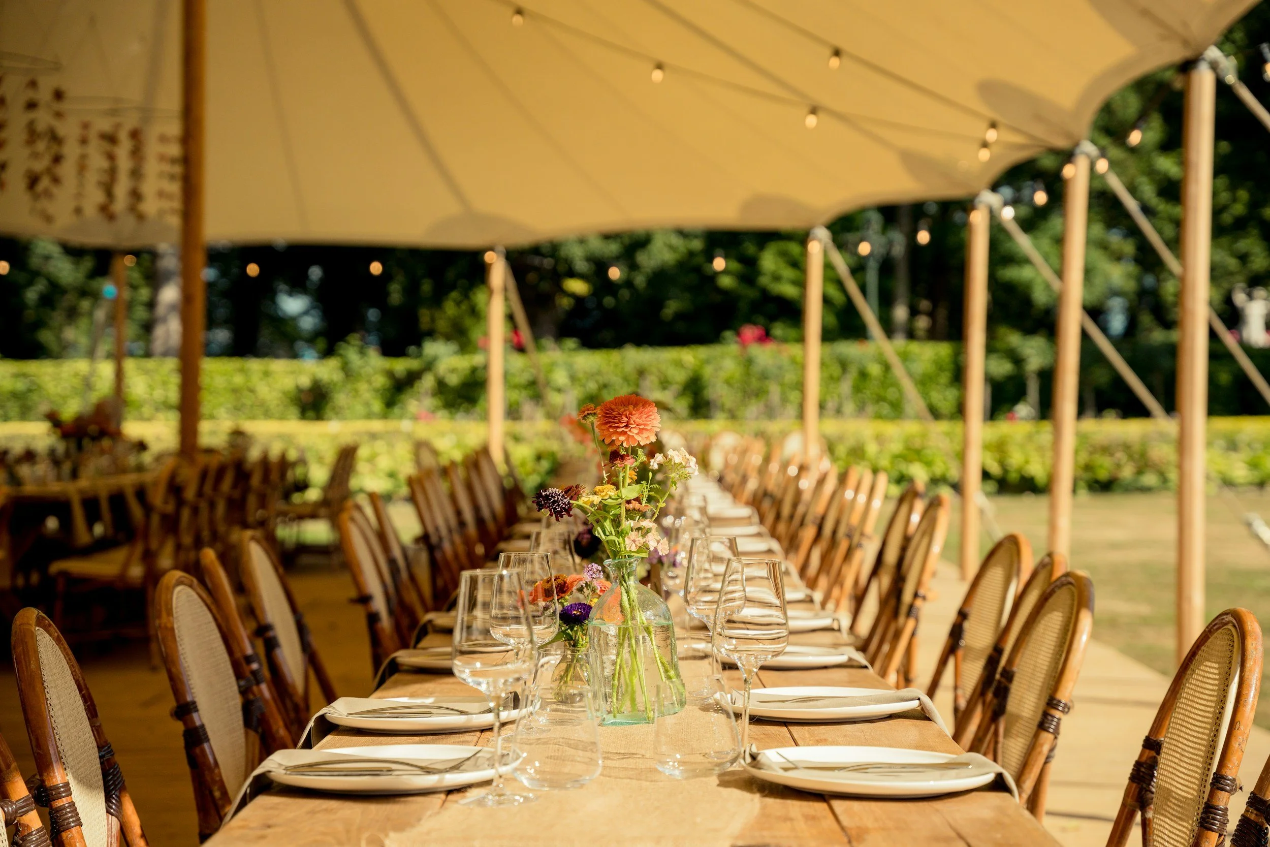 An outdoor dining table under a large canopy with flowers in the center, set with plates, napkins, and glasses, surrounded by wooden chairs.