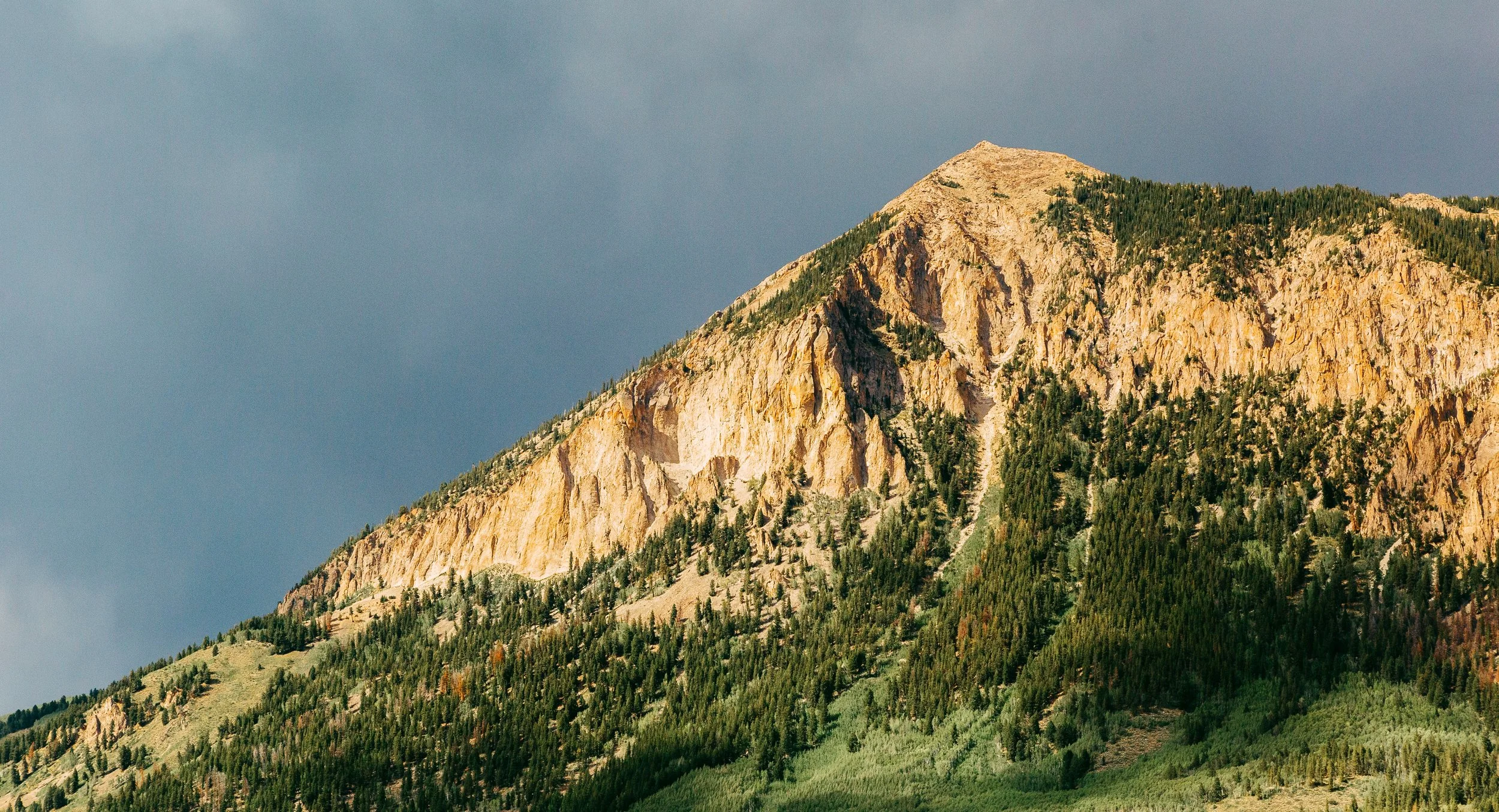 A mountain with steep rocky cliffs and green forested slopes under a cloudy sky.