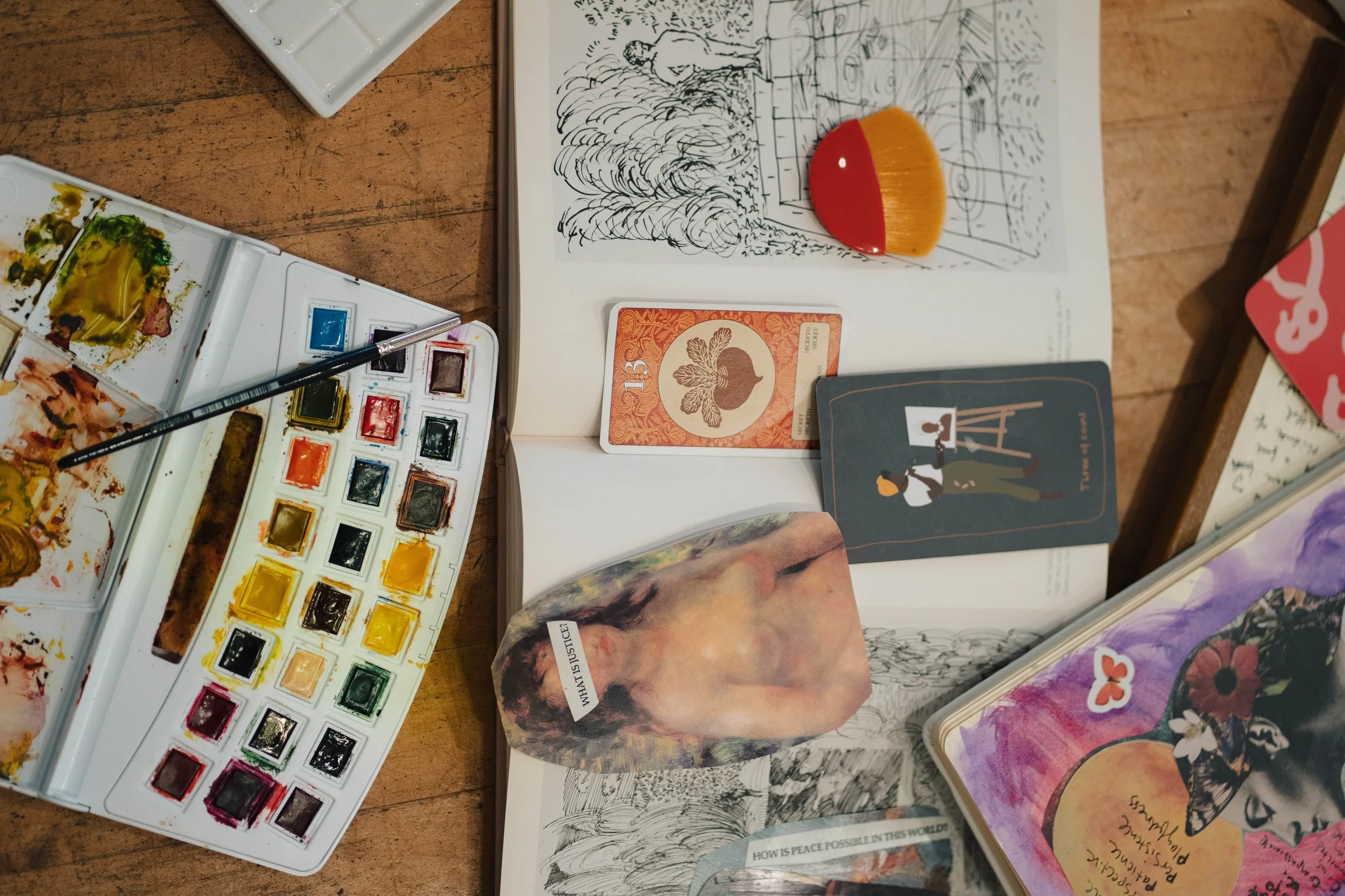 A close up of art supplies, featuring a collaged image, a messy paint palette and warm lighting.