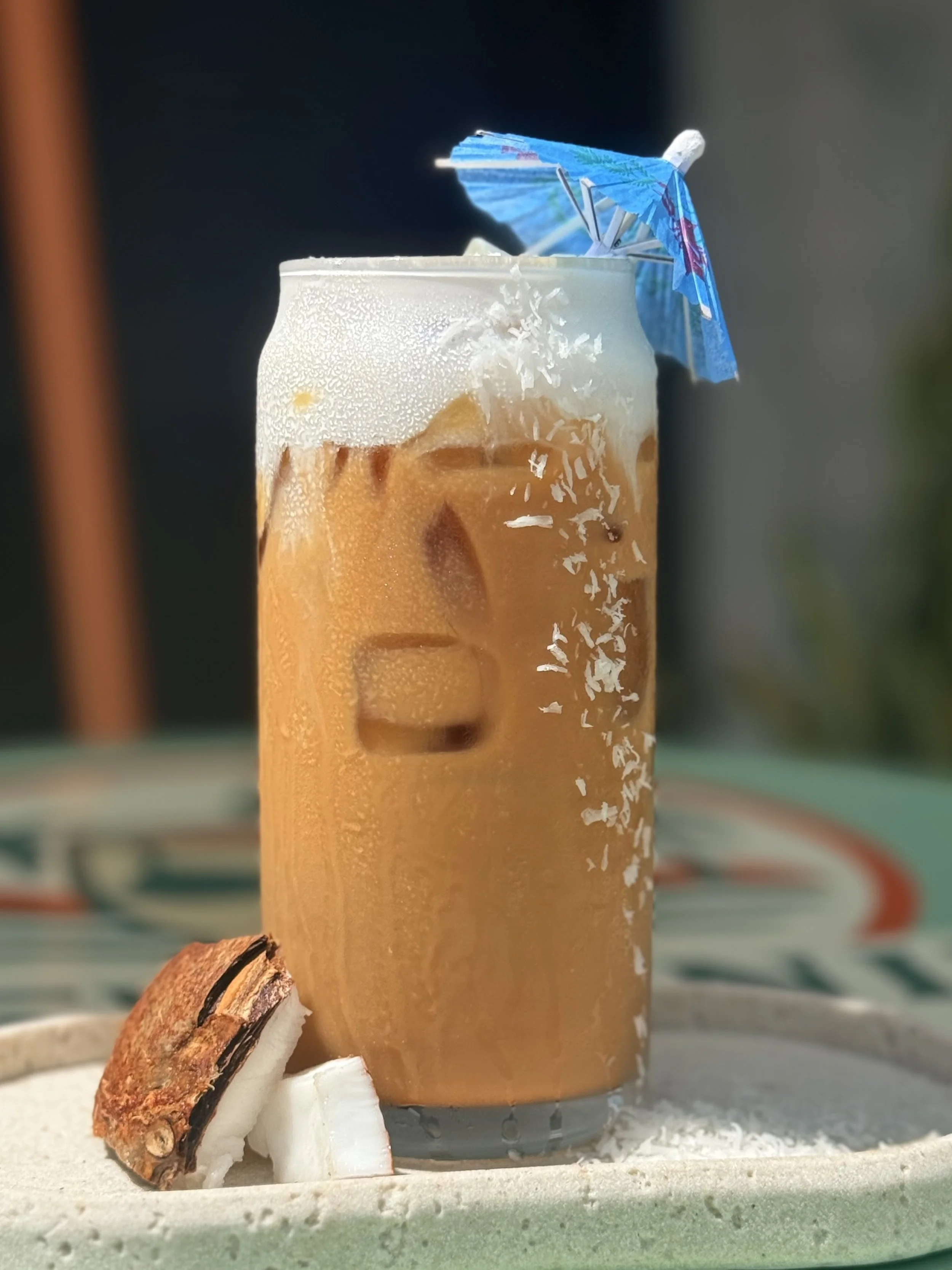 a glass with an espresso, coconut, white chocolate latte topped with cold foamwith a umbrella in it
