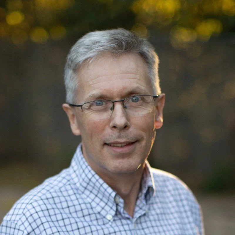 A middle-aged man with gray hair and glasses, wearing a checkered collared shirt, standing outdoors with blurred background greenery.