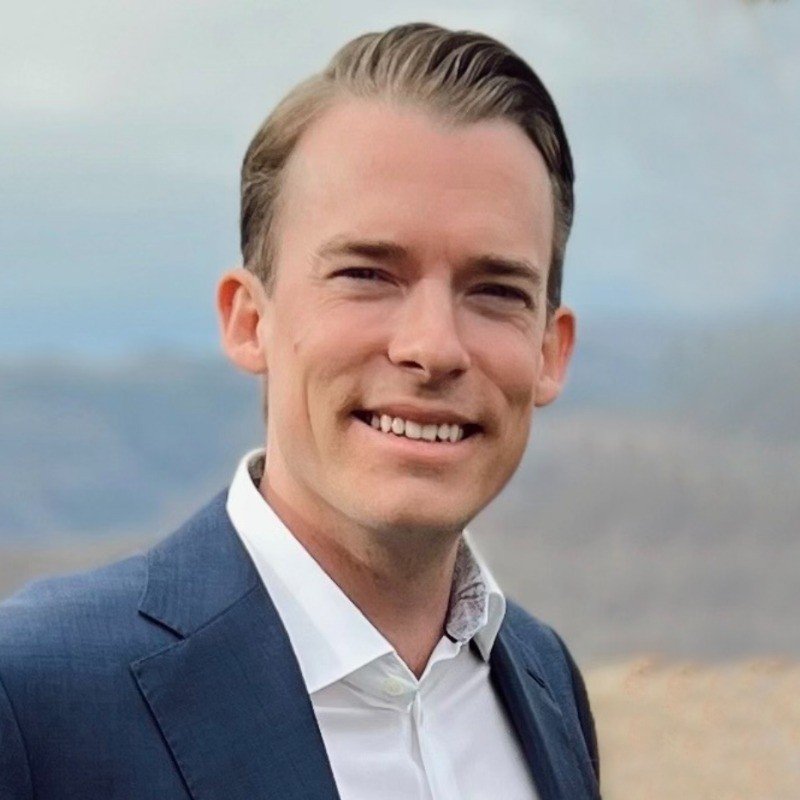 A young man with slicked-back brown hair, wearing a navy blazer and white shirt, smiling outdoors against a cloudy sky and landscape background.