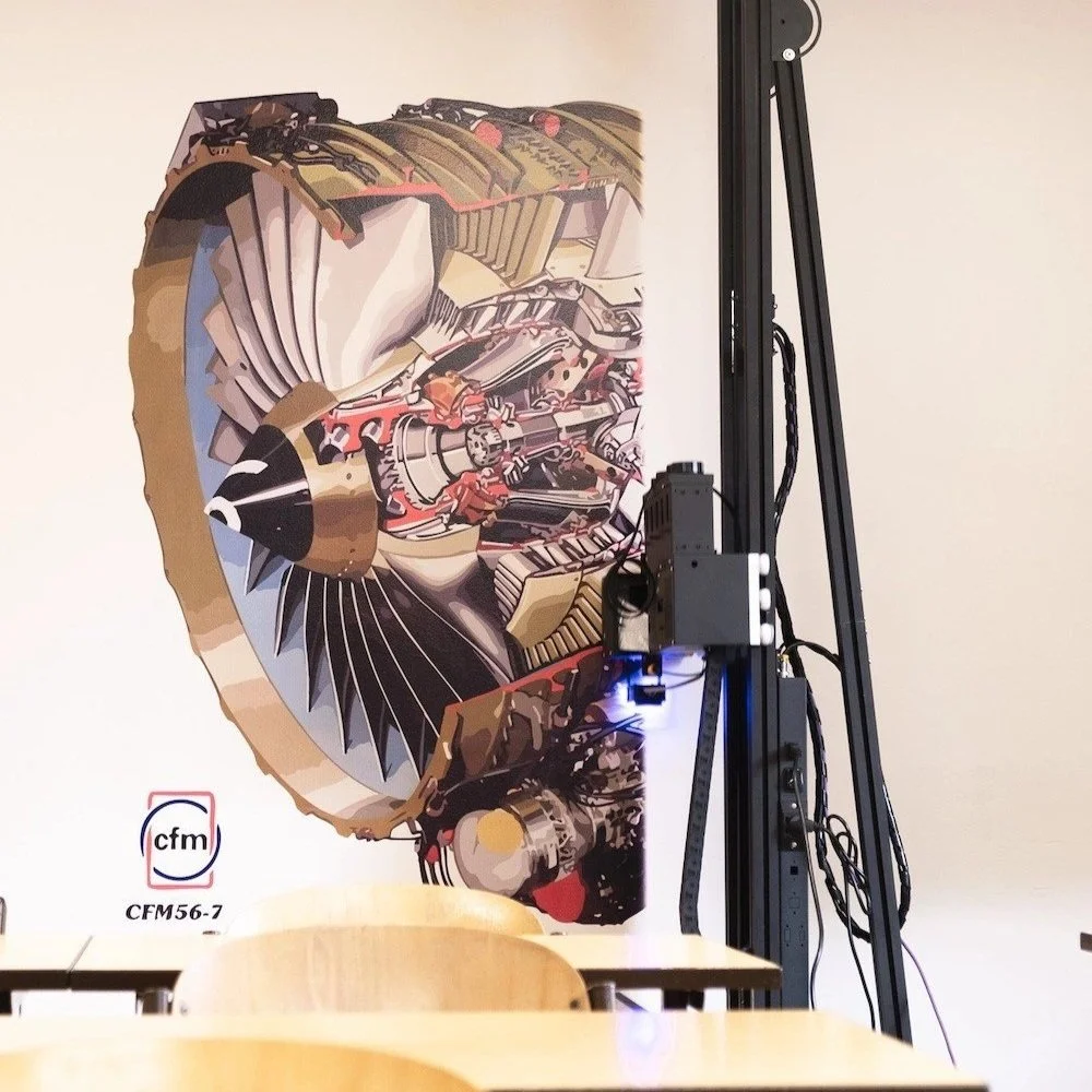 A large, colorful mural of a detailed airplane engine on a white wall, with a black robotic arm and a table in the foreground.