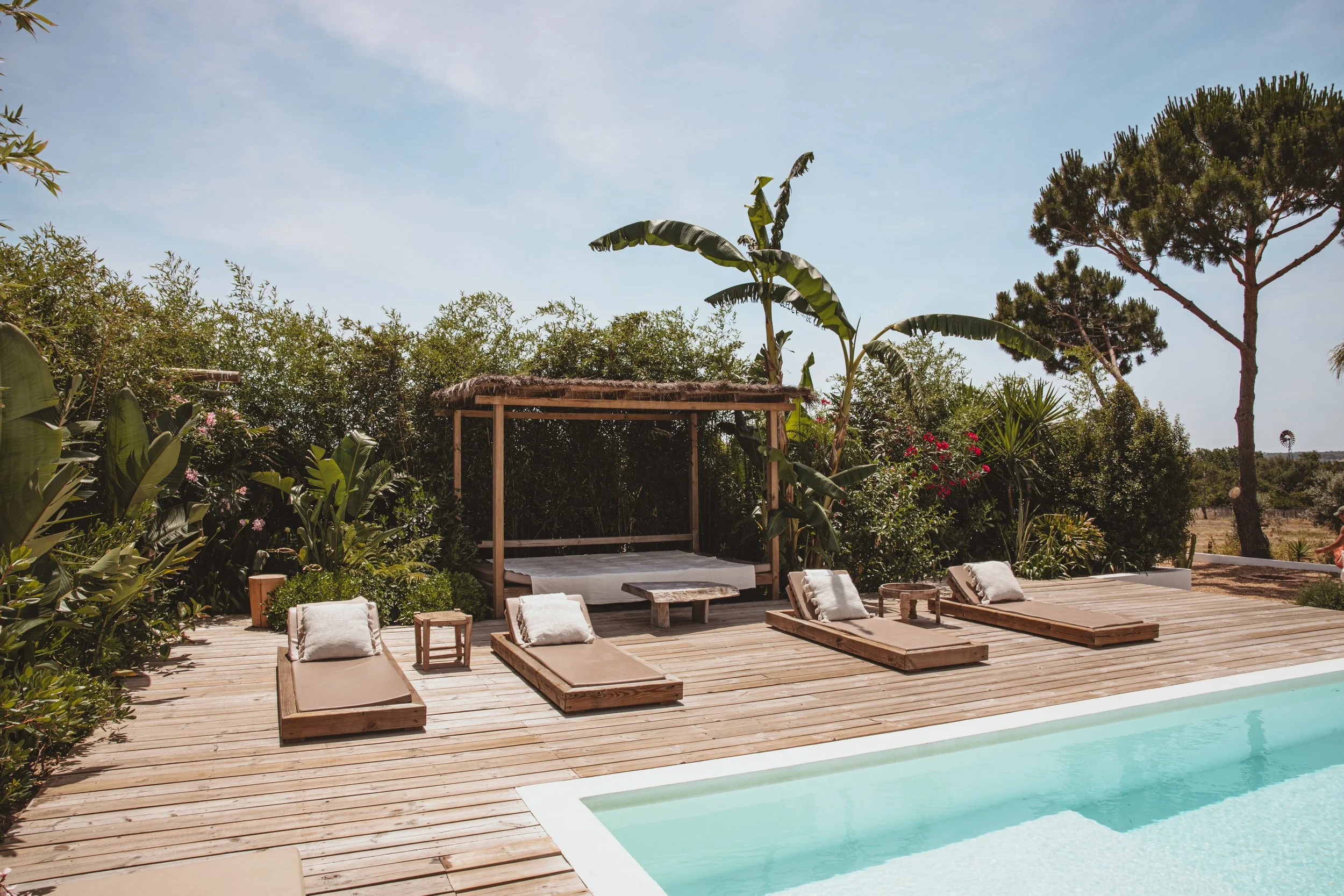 View of a backyard pool area with lounge chairs, umbrellas, greenery, a Jeep, and a bicycle, seen from inside a house through an open sliding door.