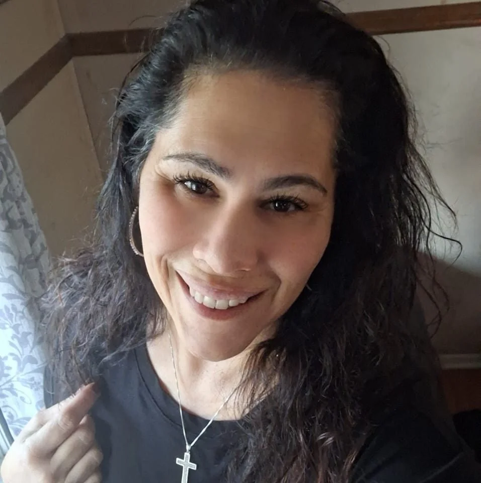 A woman with curly dark hair, smiling, wearing hoop earrings, a cross necklace, and a black shirt, standing indoors near a window with patterned curtains.