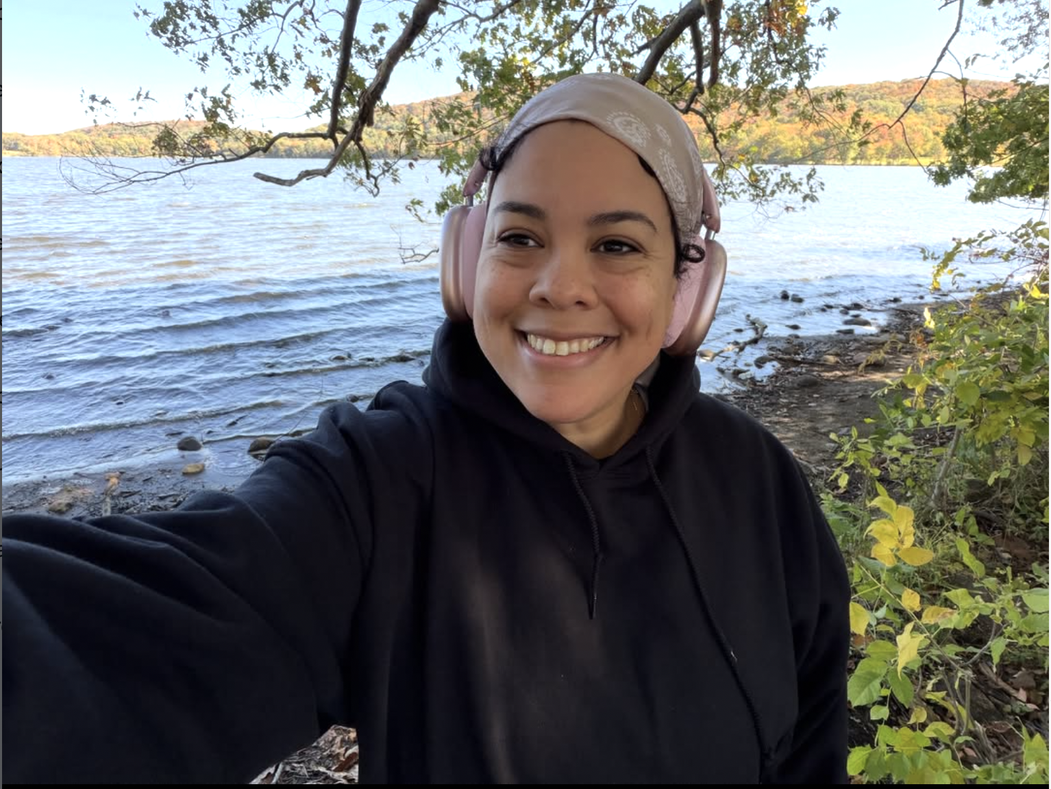 A woman smiling, wearing pink headphones and a black hoodie, taking a selfie outdoors near a lake with trees and mountains in the background.