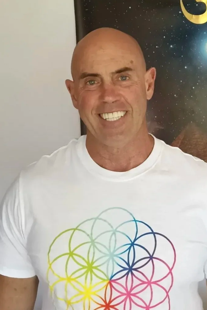 A smiling man with a bald head and light complexion, wearing a white t-shirt with a colorful geometric flower of life design, standing in front of a space-themed poster with a crescent moon and stars.