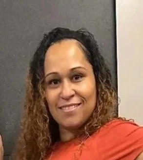 A woman with curly hair smiling in front of a gray and white background.
