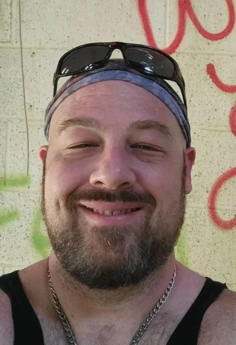 Close-up of a smiling man wearing sunglasses on his head, a headband, and a black tank top, standing against a graffiti-covered wall.