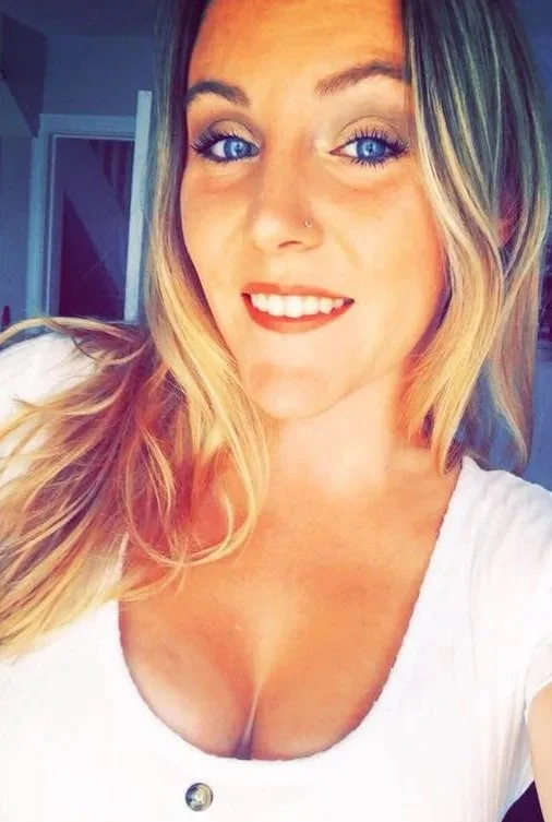 A young woman with long blonde hair and blue eyes taking a selfie indoors, smiling with red lipstick, wearing a white shirt with a button, and standing in front of a door and walls painted in a neutral color.