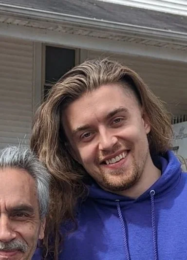 A smiling young man with long, wavy hair wearing a blue hoodie, standing beside an older man with gray hair, both outdoors near a house.