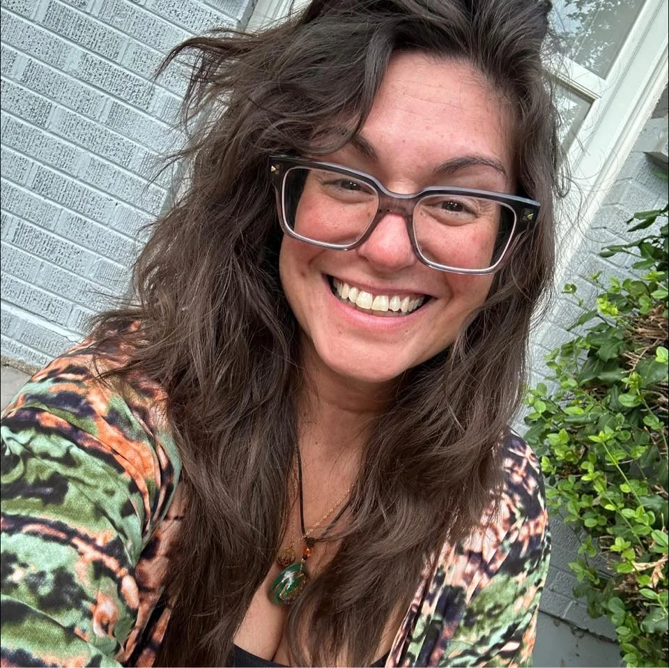 A smiling woman with wavy brown hair wearing large glasses and a colorful patterned top, standing outdoors near a gray brick wall and green bushes.