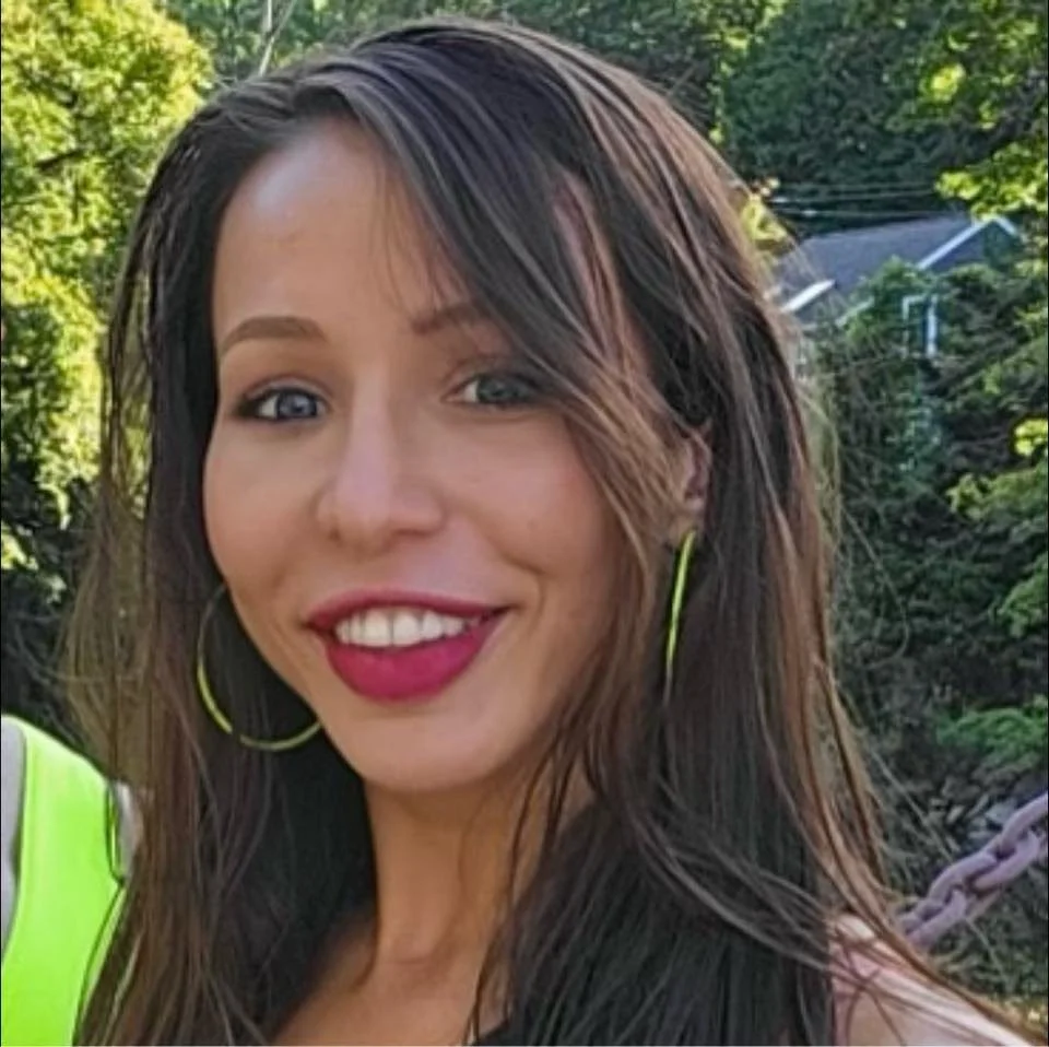 A woman with long brown hair, blue eyes, wearing makeup including bright lipstick, hoop earrings, and a high-visibility yellow vest, smiling outdoors with green trees and a house in the background.