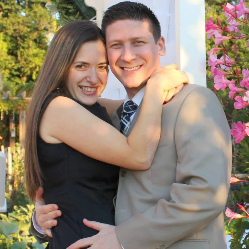 A smiling young man and woman hugging outdoors with trees and pink flowers in the background.