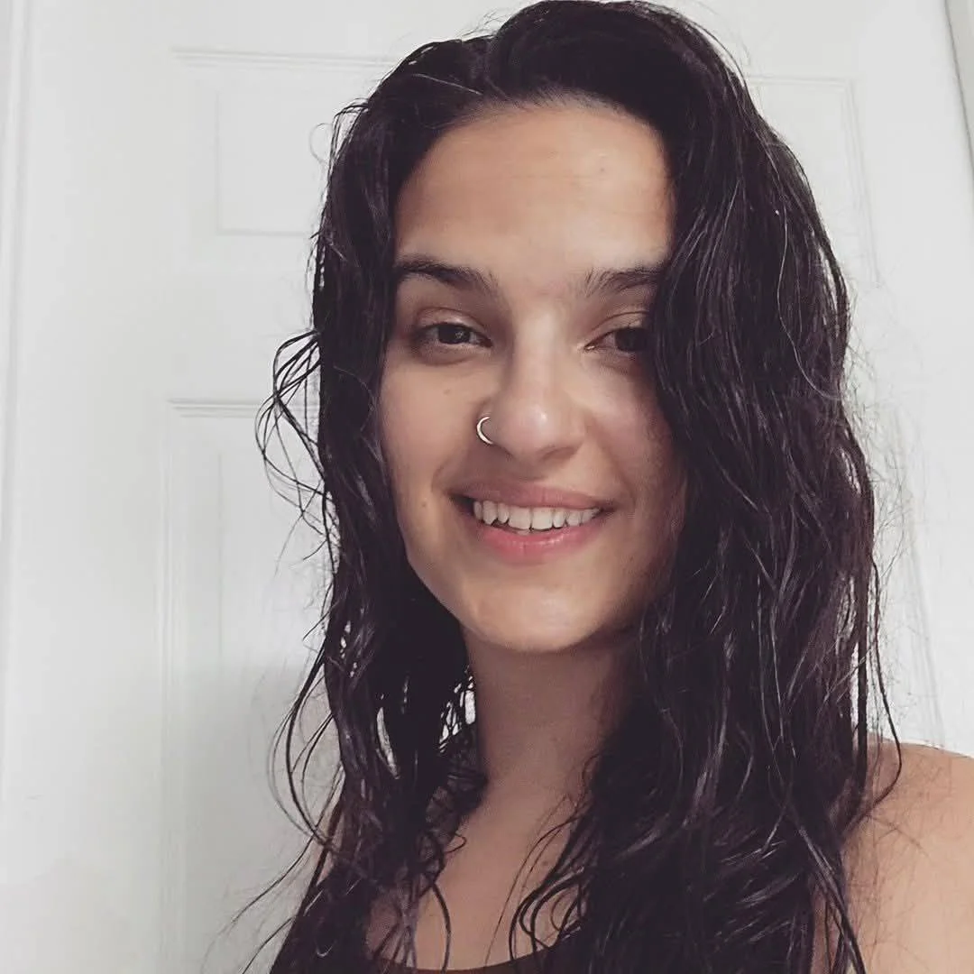 A woman with long curly dark hair, a nose ring, and a slight smile, standing in front of a white door.