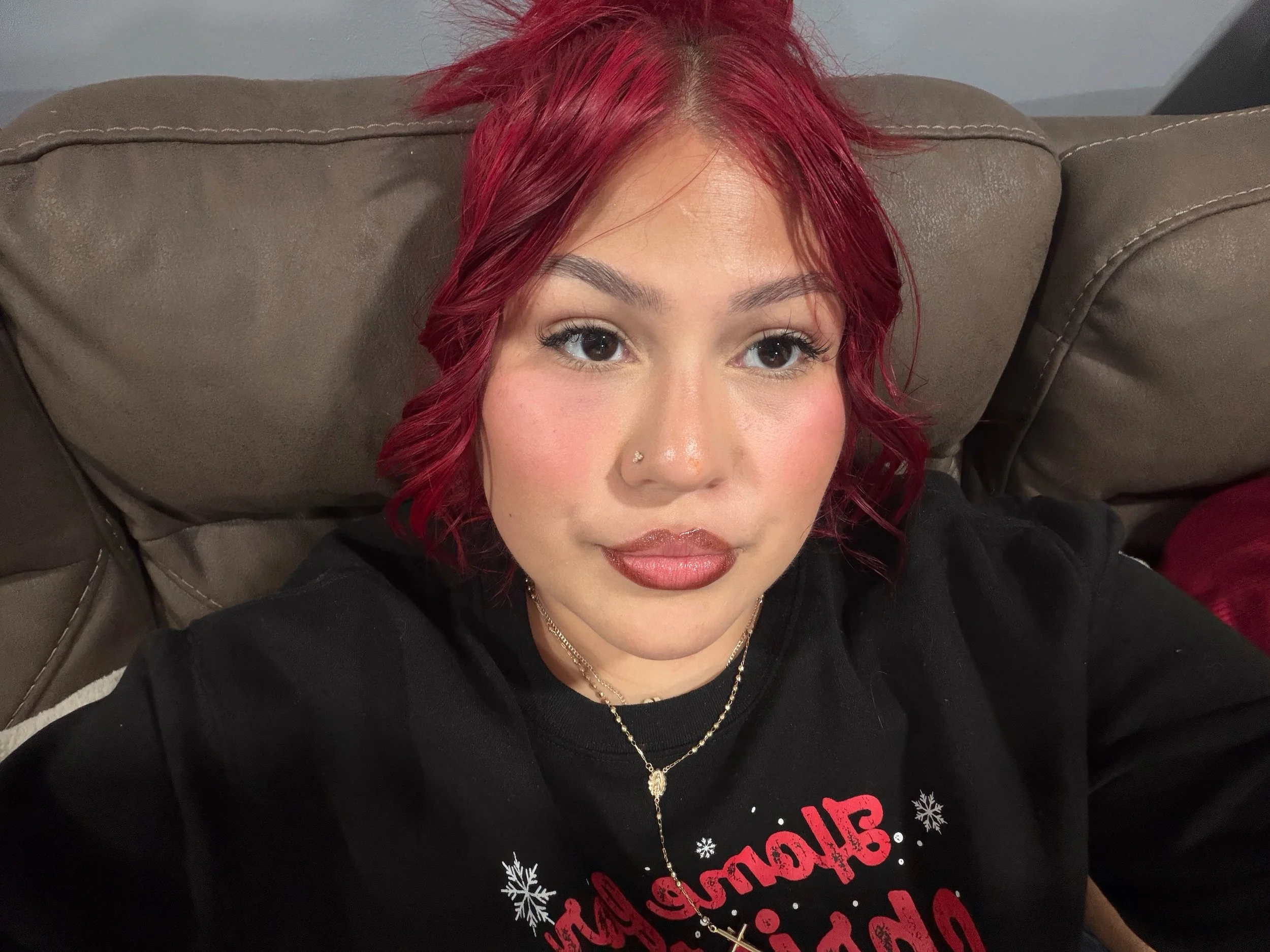 A woman with red hair and makeup taking a selfie while sitting on a brown couch, wearing a black shirt with red and white lettering.