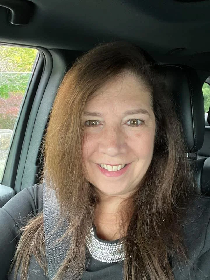A woman with long brown hair and a big smile sitting in a car, wearing a black top with a silver necklace.
