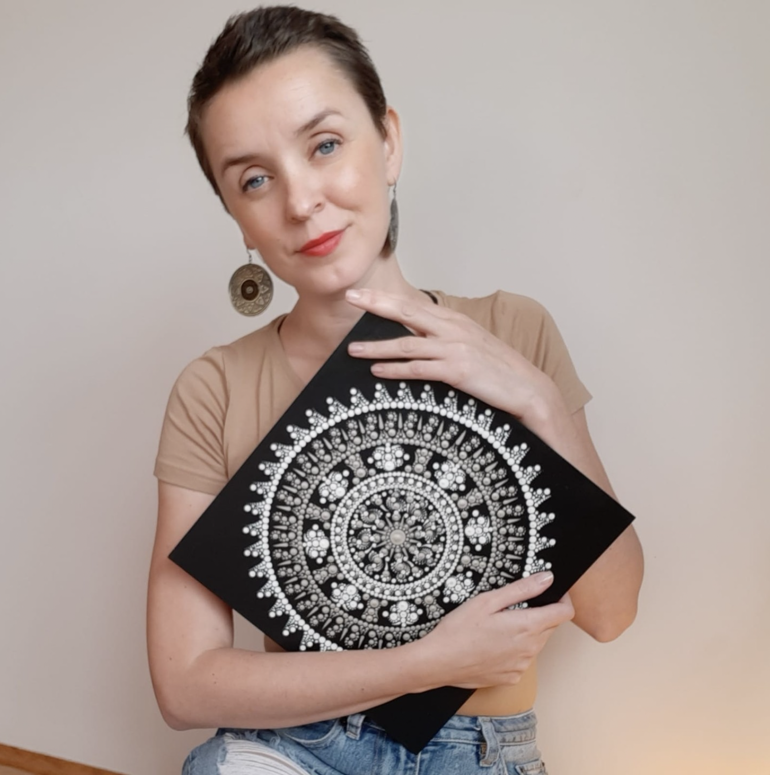 A woman with short dark hair, wearing a beige top and earrings, holding a black and white patterned artwork in front of her. She is smiling and posing against a plain beige wall.