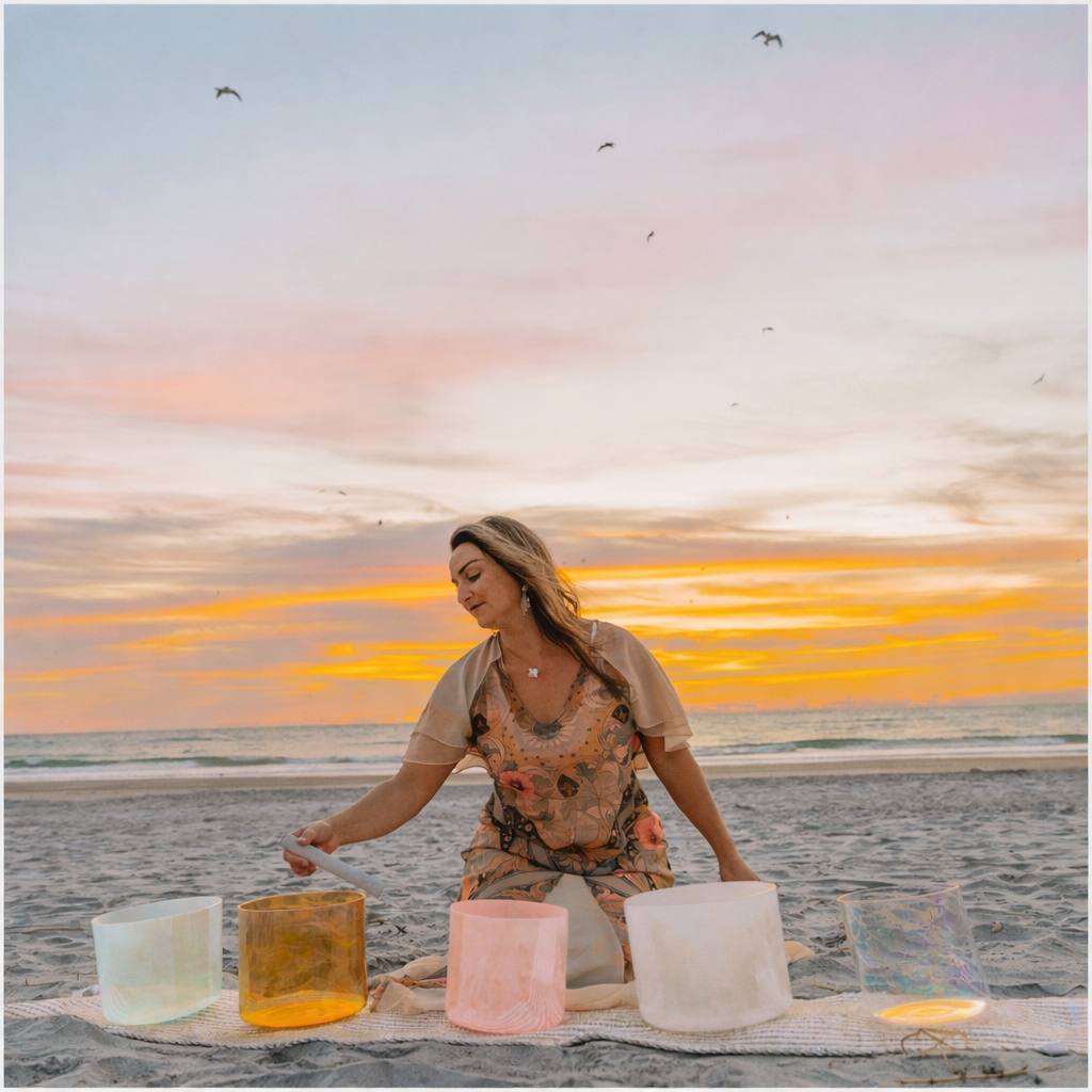 A woman playing crystal bowls on a beach at sunset, with a colorful sky and seagulls flying overhead.