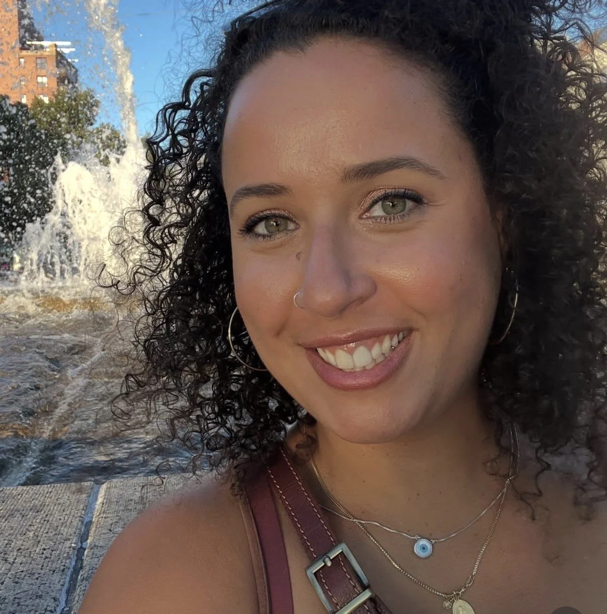 A woman smiling outdoors near a fountain with water spray, wearing hoop earrings and layered necklaces, in bright sunlight.