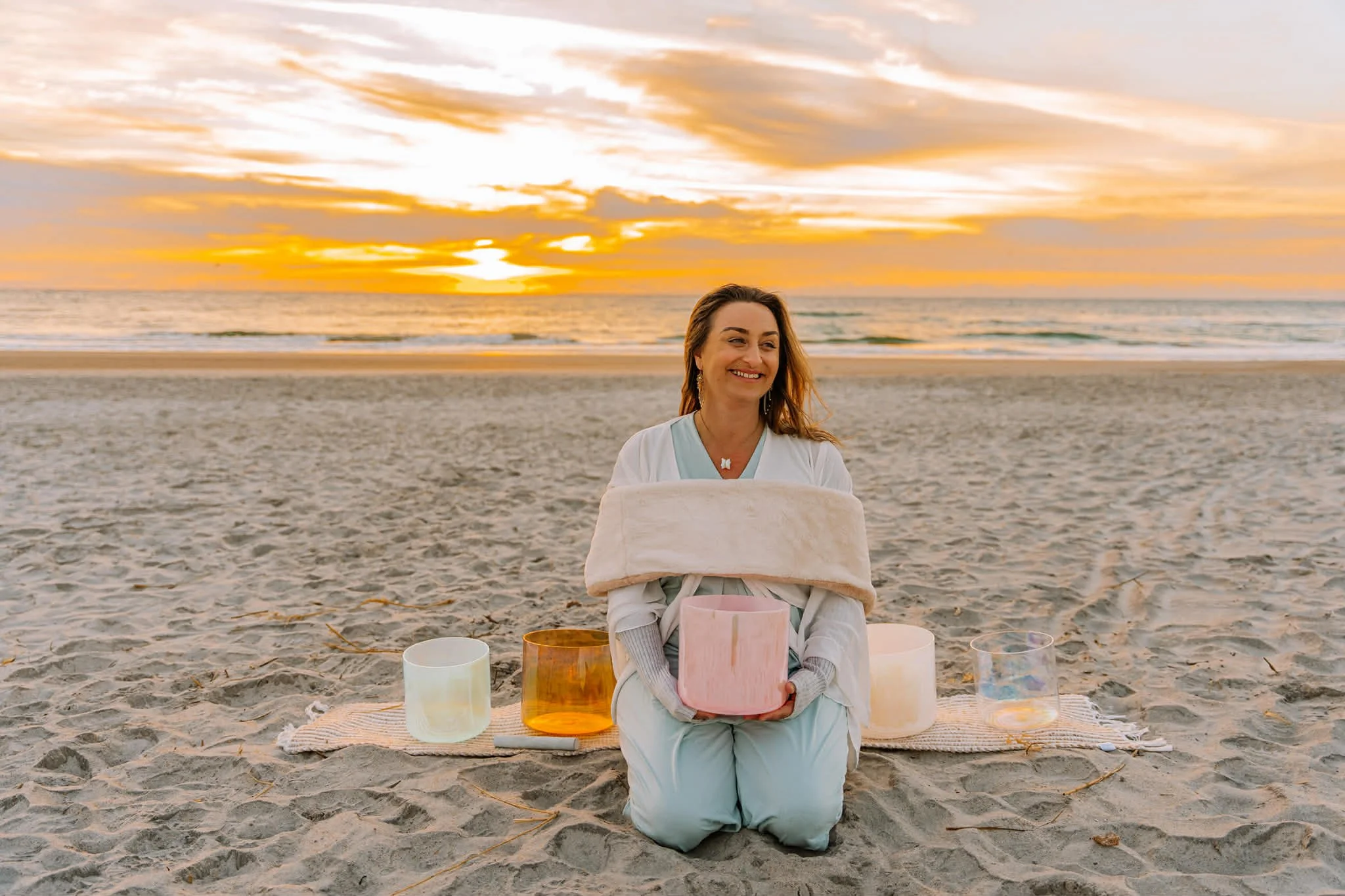 A woman sitting on the sand at the beach during sunset, holding a pink crystal singing bowl, with other crystal bowls placed on a blanket beside her, smiling and surrounded by a serene atmosphere.