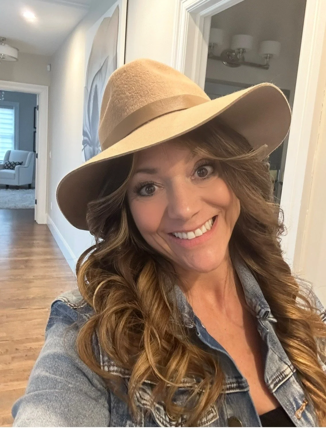 A woman smiling, wearing a large beige floppy hat and a denim jacket inside a home.