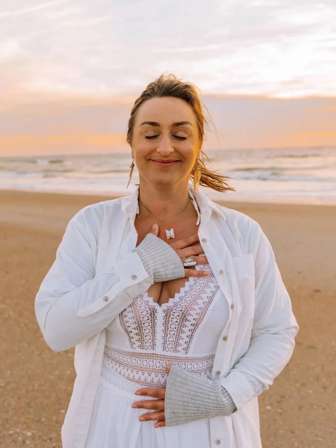 A woman with closed eyes and a peaceful smile, standing on a beach at sunset, touching her chest with her right hand.