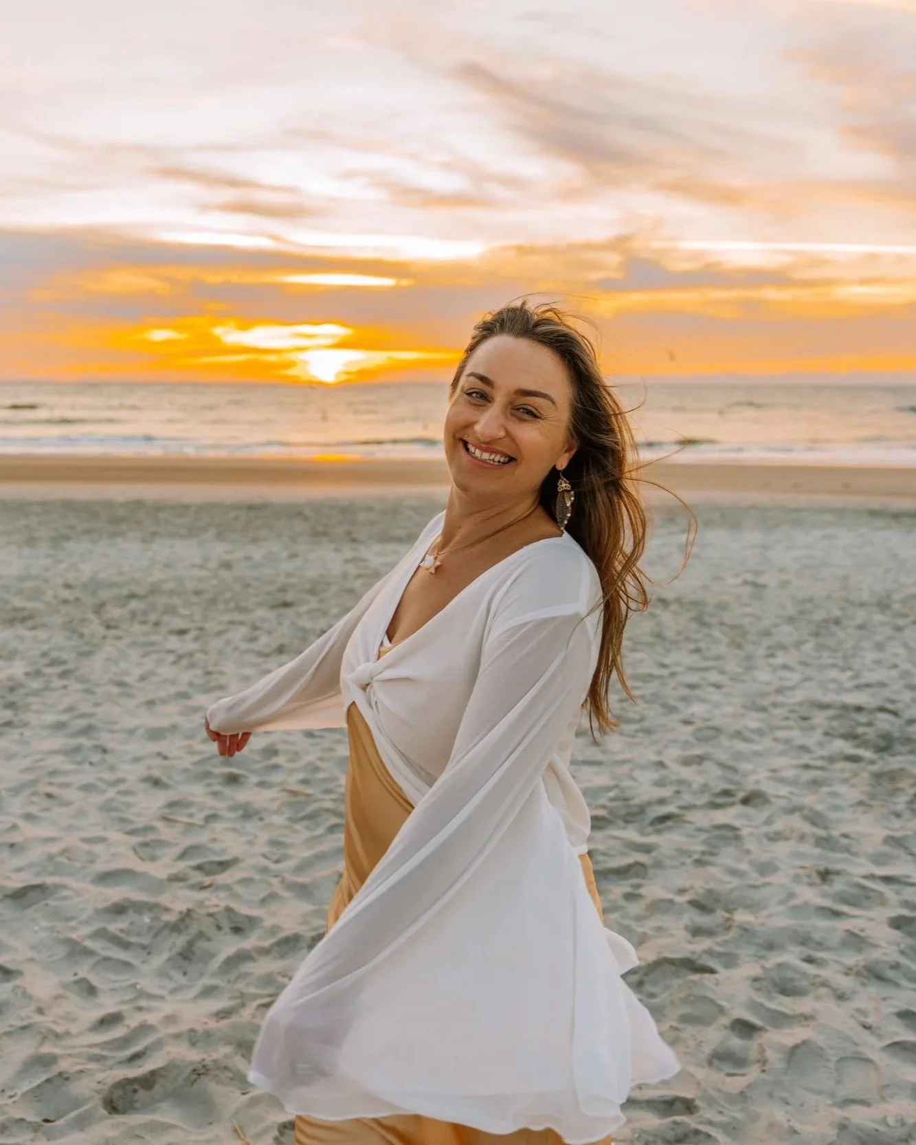 Woman smiling and wearing a white blouse and beige skirt on the beach during sunset.