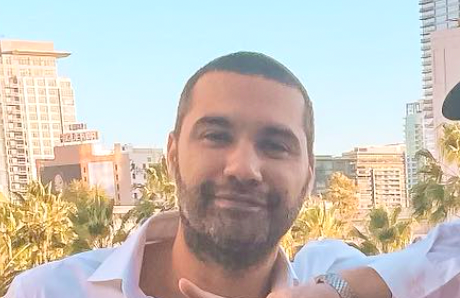 Man with short dark hair and beard smiling outdoors with city buildings and palm trees in the background.