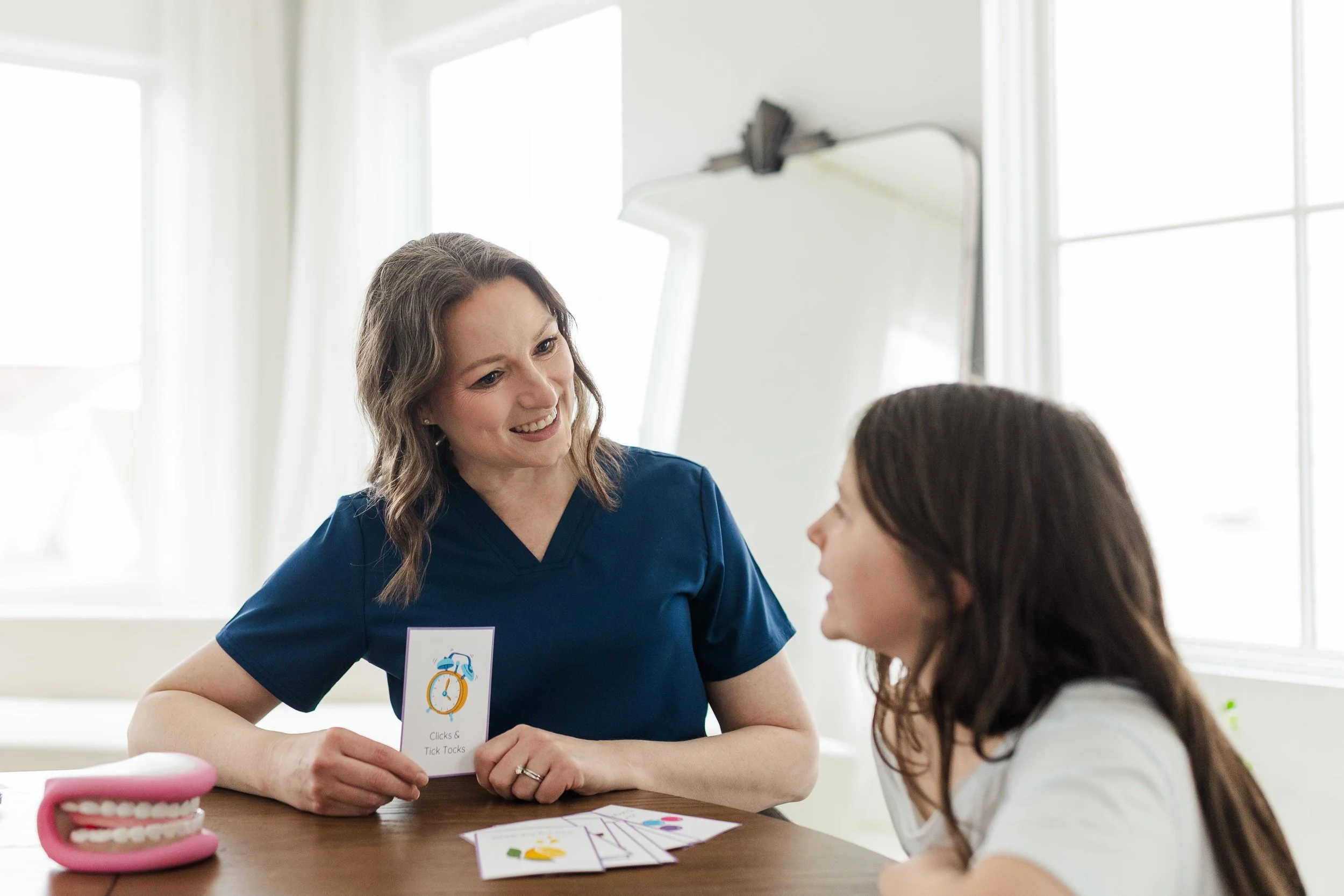 A woman in blue scrubs teaching a young girl a language game at a table in a well-lit room.