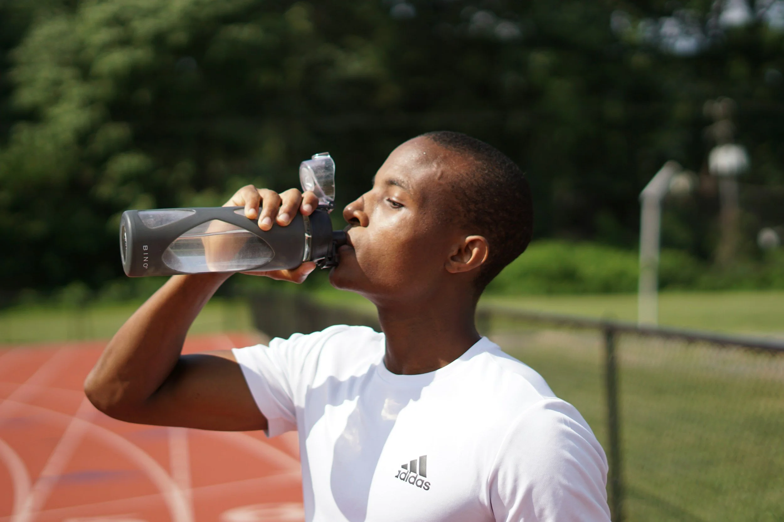 A young man in a white Adidas shirt drinking water from a clear plastic bottle at an outdoor running track.