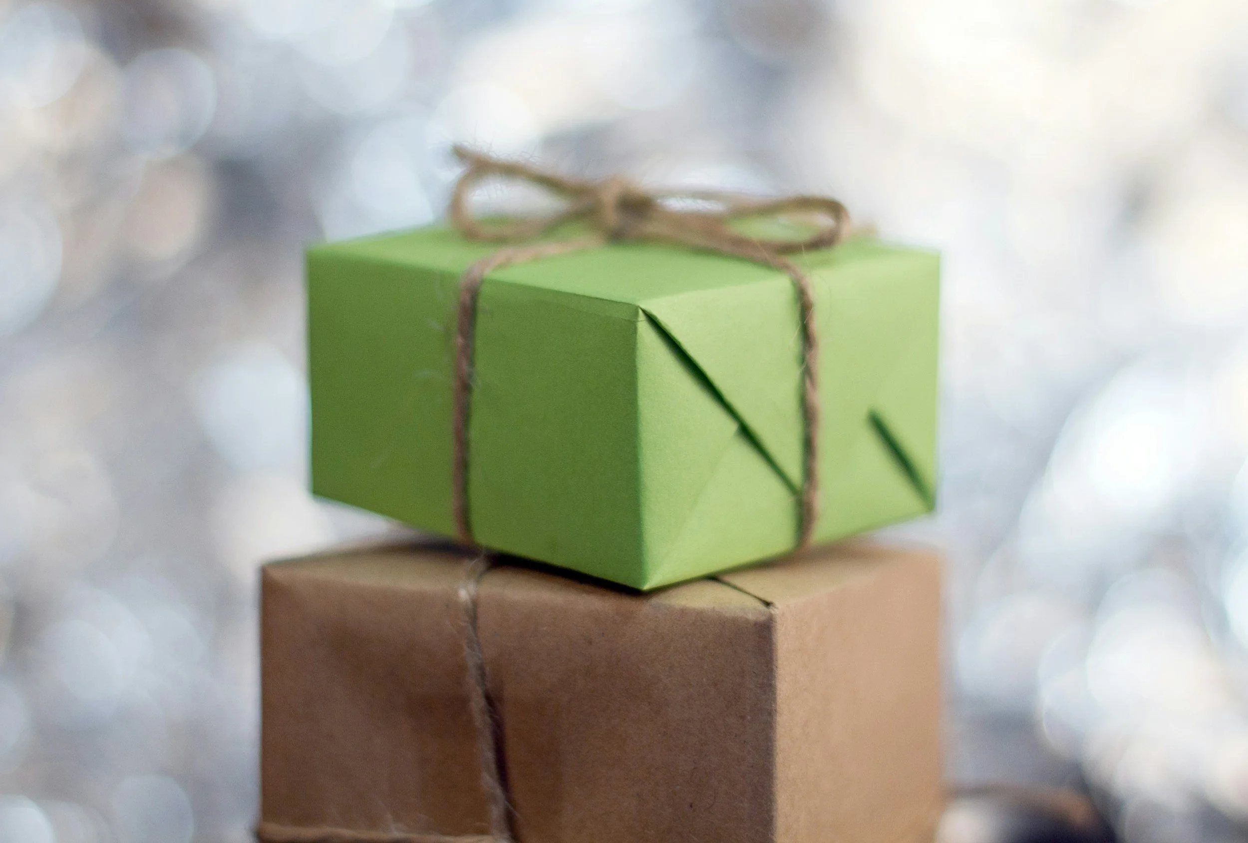 A small green gift box with a brown string tied in a bow, sitting on top of a larger brown gift box, with a blurred, sparkly background.