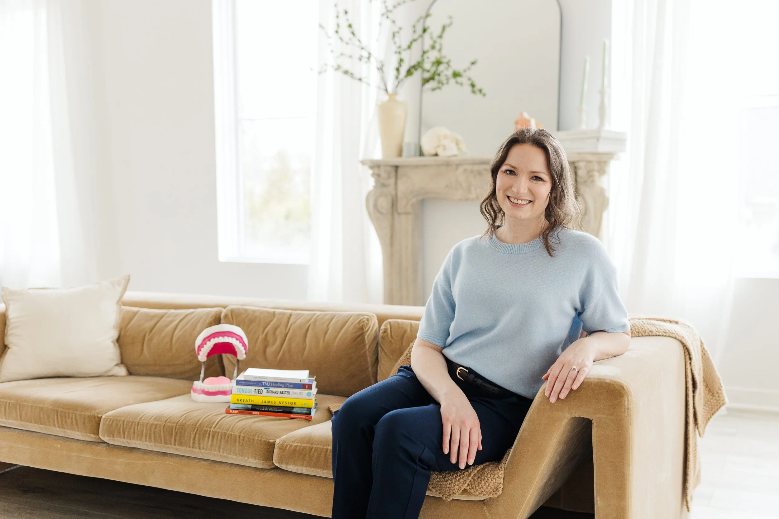 Woman sitting on beige sofa in bright living room, smiling, with a stack of books and a dental model