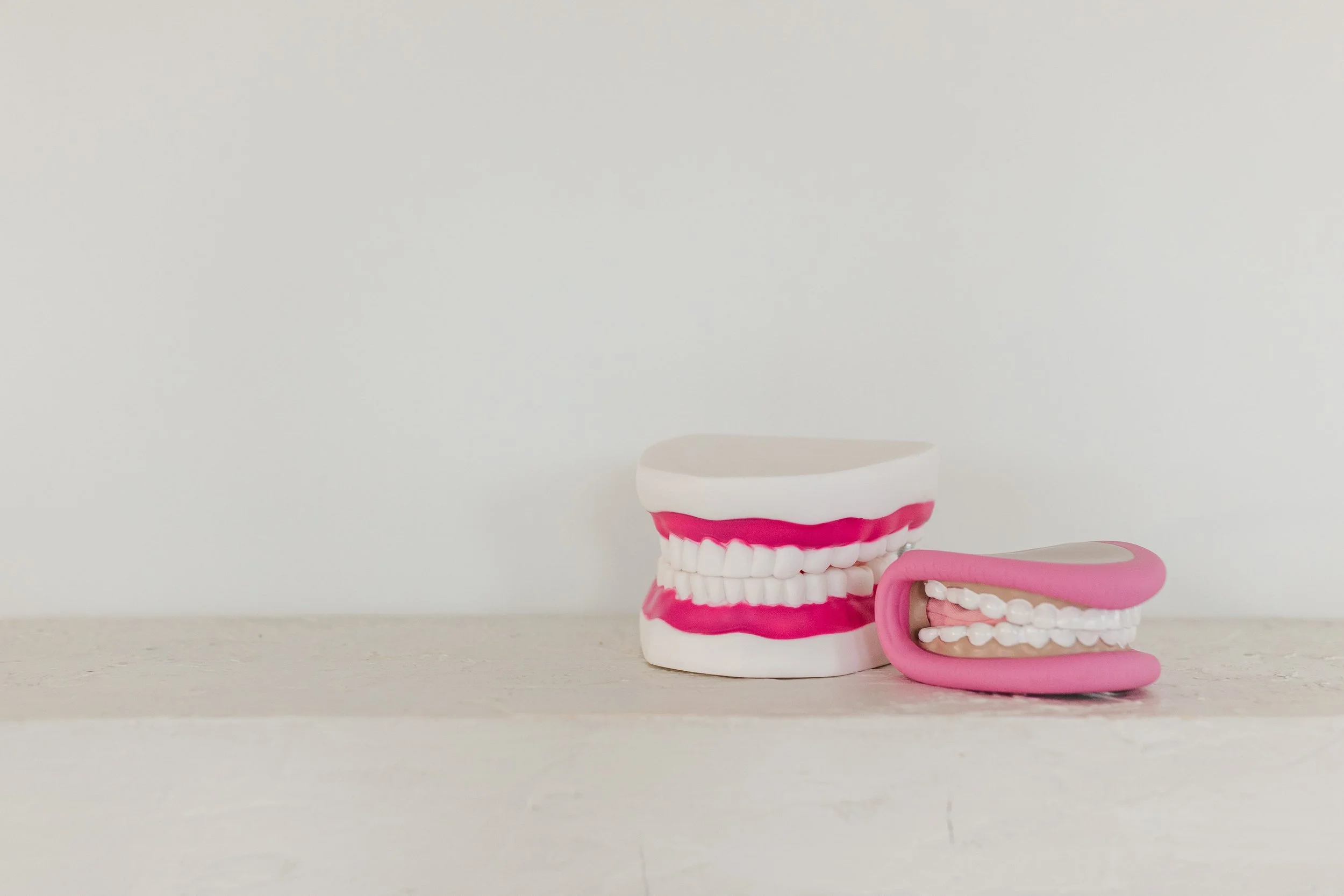 A dental model with white teeth and pink gums next to a pink gum model on a white surface against a plain white wall.